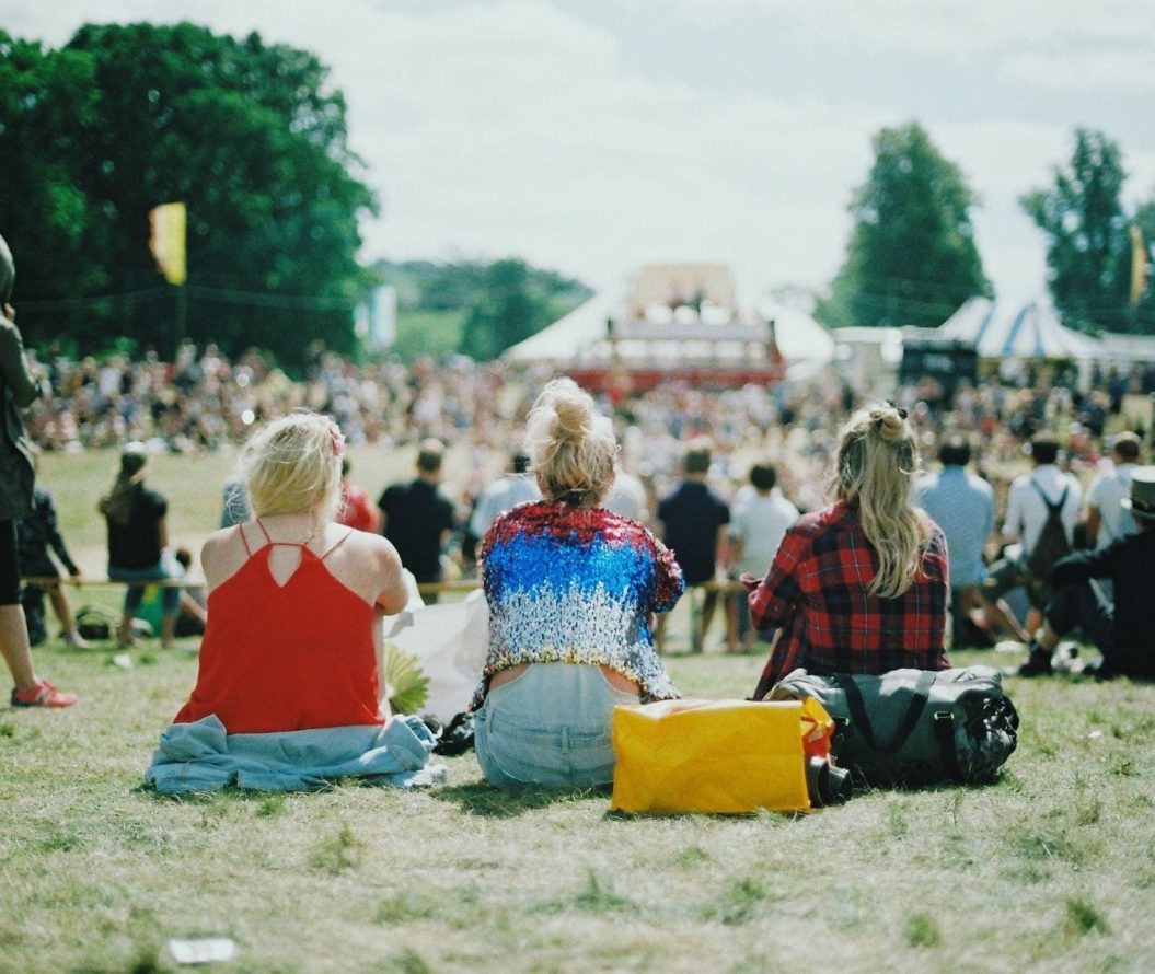 The back of three girls sat facing a stage at a festival.