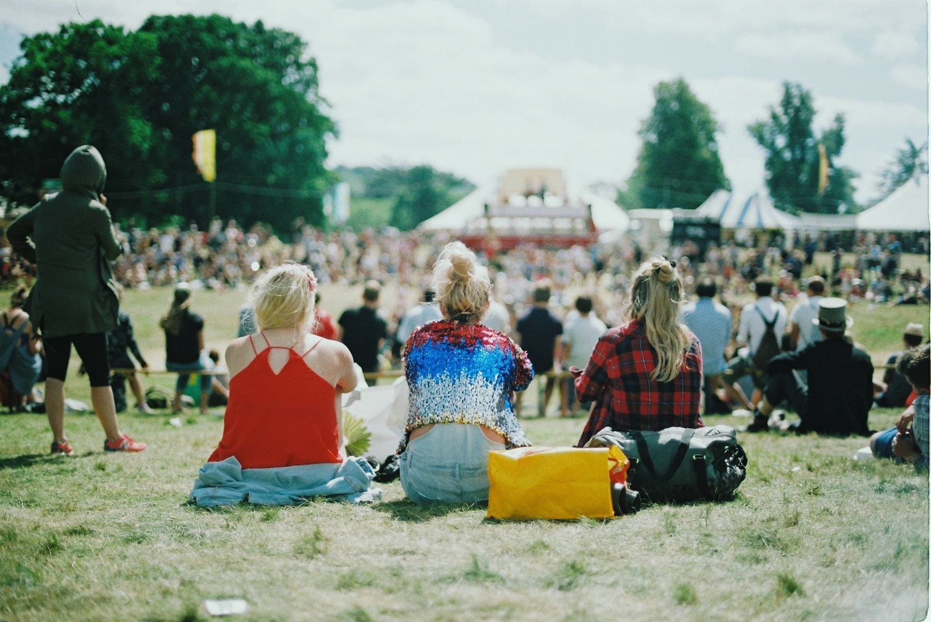 The back of three girls sat facing a stage at a festival.