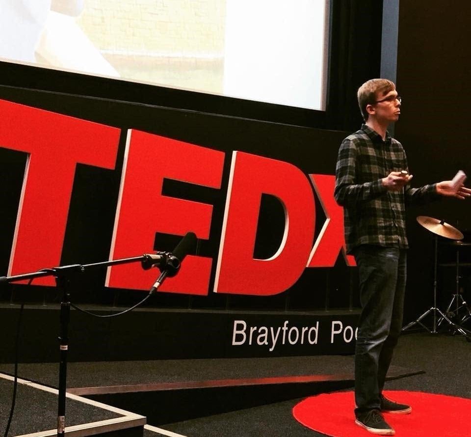 Male student standing in front of a TedX sign, delivering a speech.