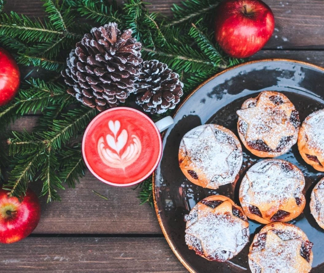 mince pies on a plate and a hot, red drink in a mug. There are decorative apples and pinecones surrounding them.