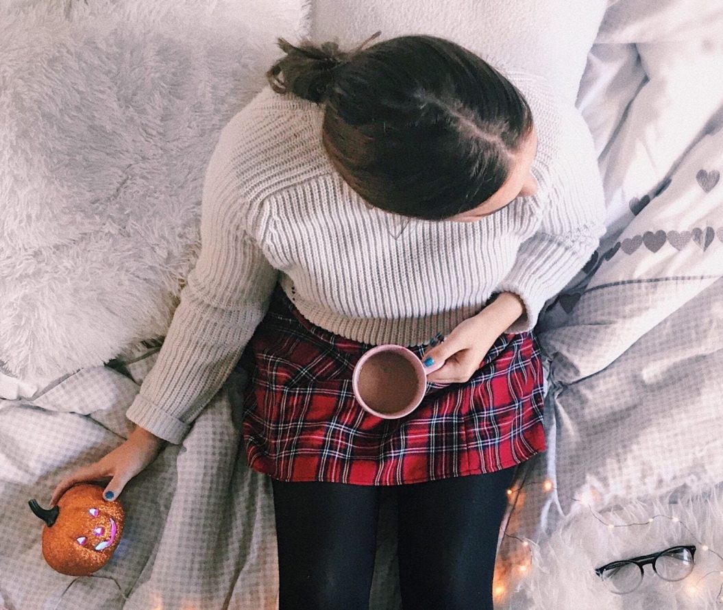 A young woman sits on a white bed whilst holding a hot chocolate and a Halloween pumpkin decoration