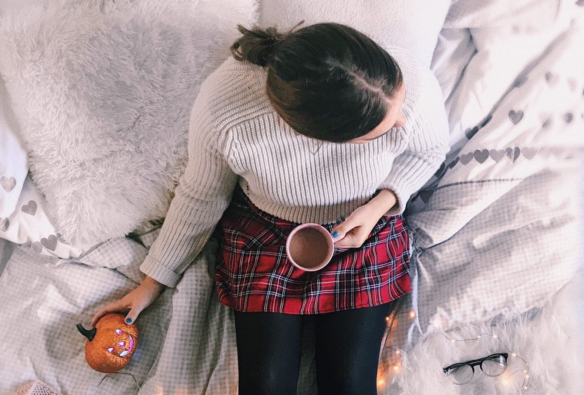 A young woman sits on a white bed whilst holding a hot chocolate and a Halloween pumpkin decoration