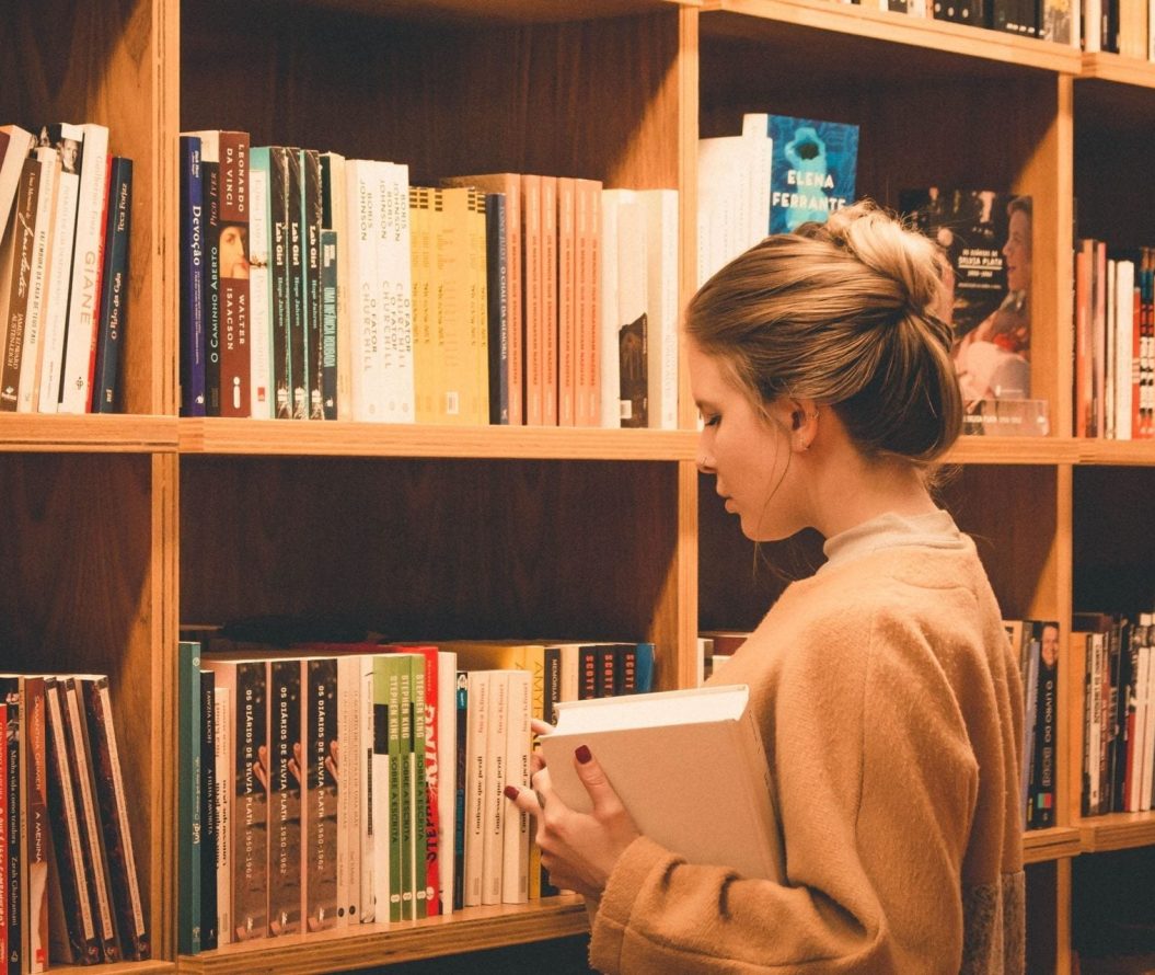 A girl stood in front of a bookshelf full of books, holding a book