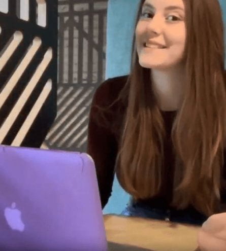A girl smiling sat at a desk in front of a laptop
