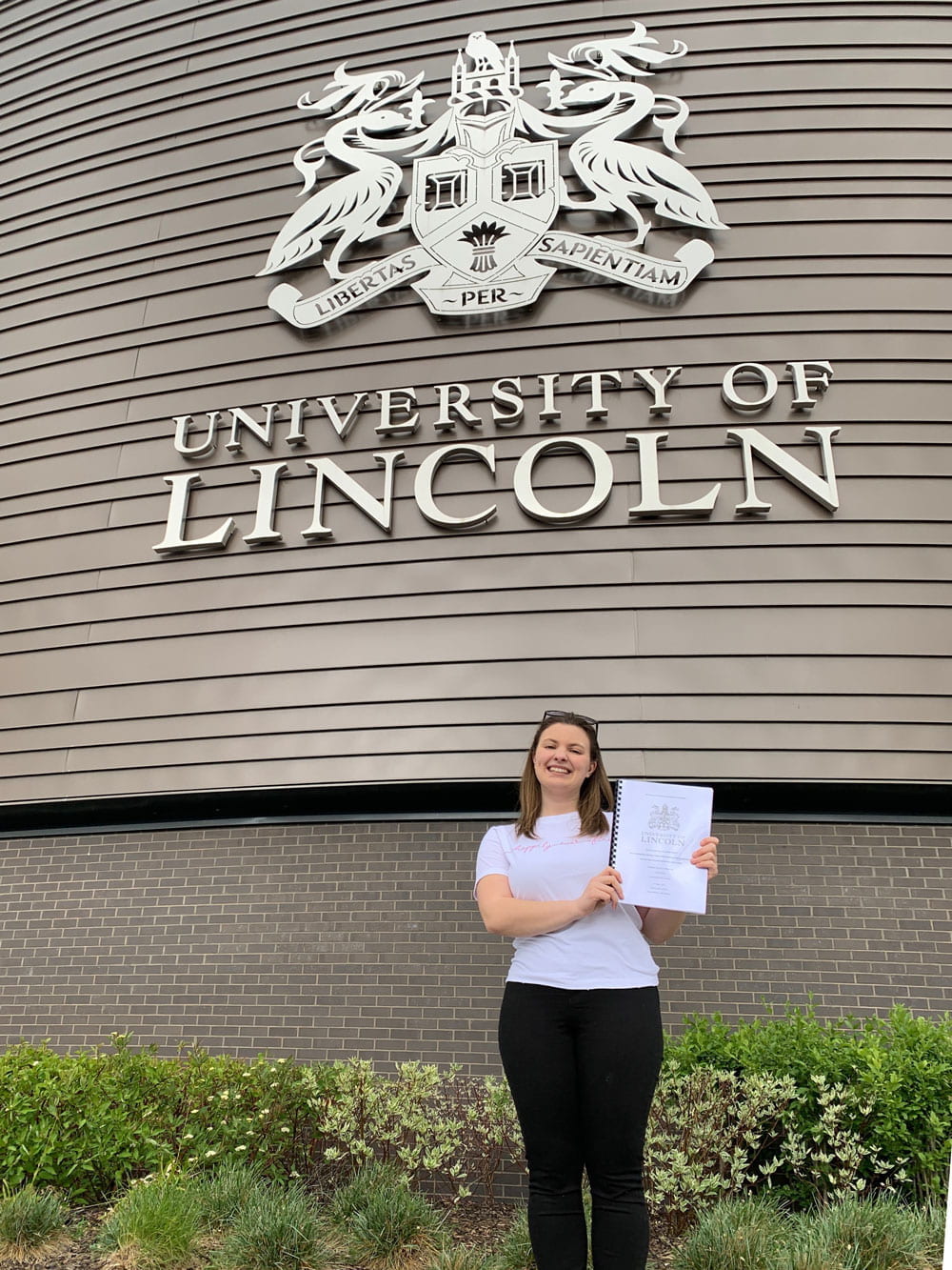 A young woman holding a dissertation and smiling in front of the University of Lincoln logo on a building