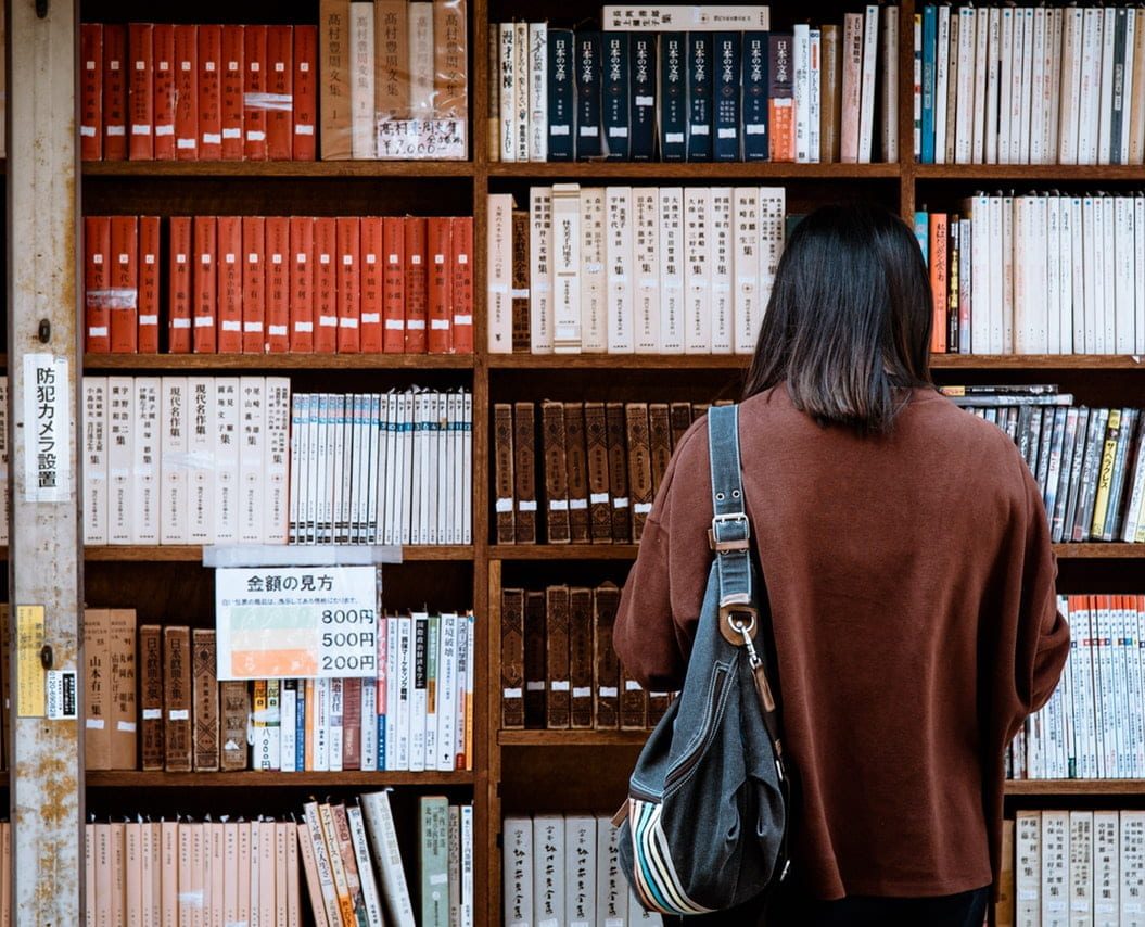 A woman wearing a brown shirt stands in front of a bookcase full of books