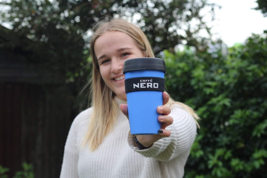 A girl holding a blue Caffe Nero cup towards the camera in a garden.