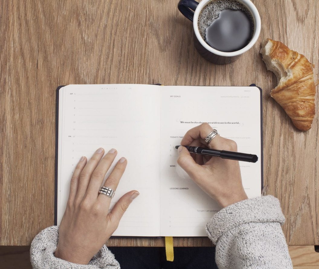 a woman wearing rings, eating a croissant and having a coffee writing in her planner