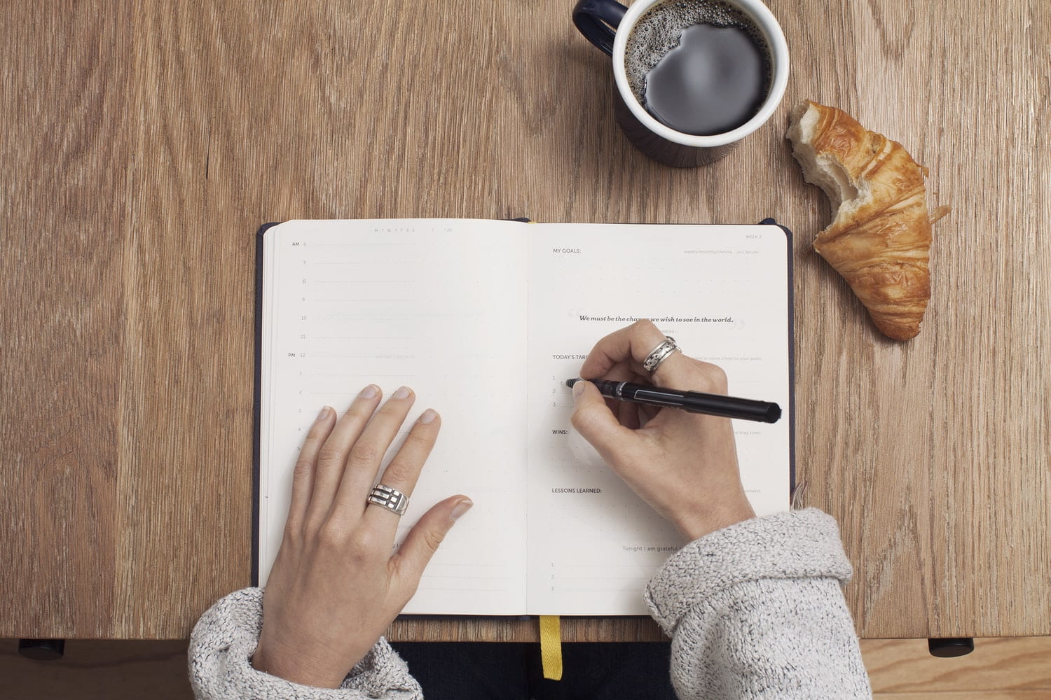a woman wearing rings, eating a croissant and having a coffee writing in her planner