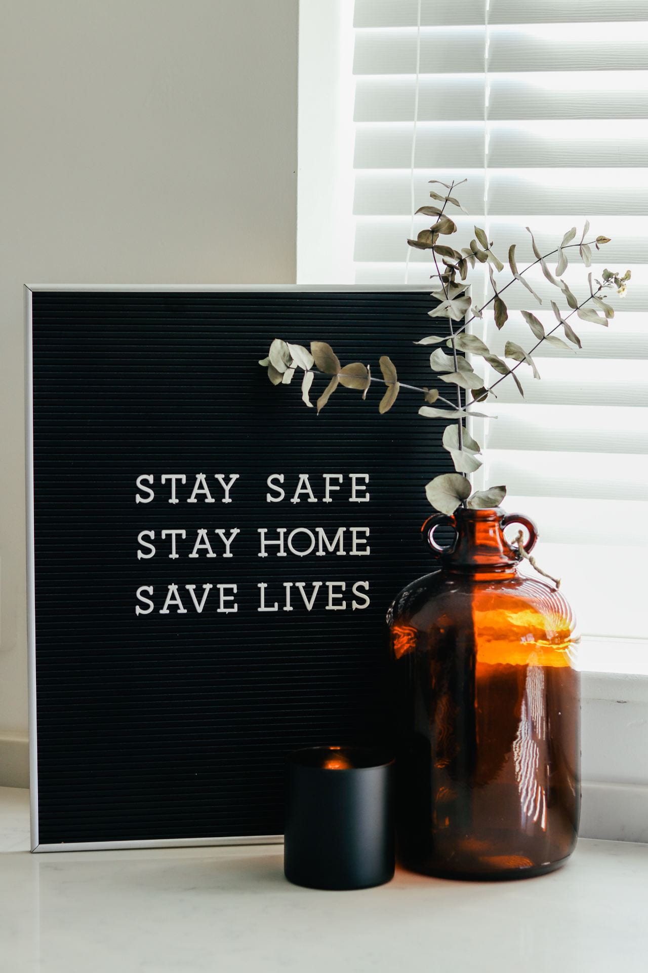 Framed photograph that reads: Stay Safe, Stay Home, Save Lives. Potted plant and a candle are in the foreground of the image.