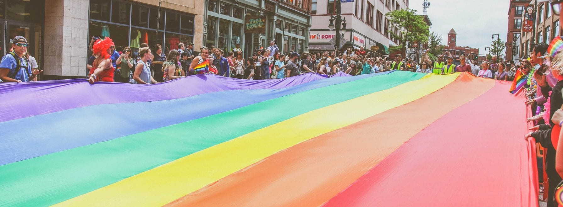 large rainbow flag being held by a large group of people in a city centre