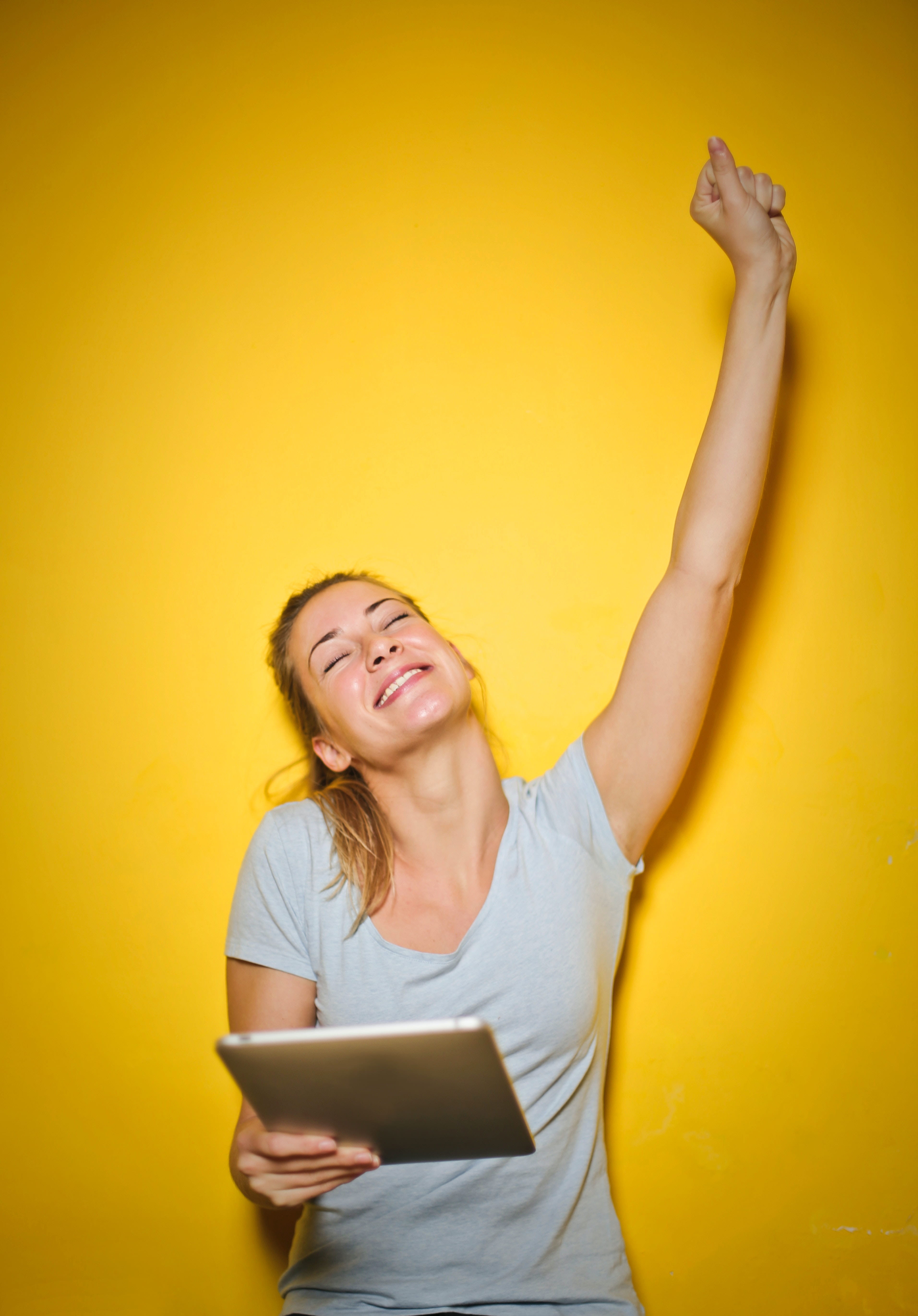 A woman holding an iPad in one hand with the other arm raised into a fist upwards. She is smiling. There is a yellow background.