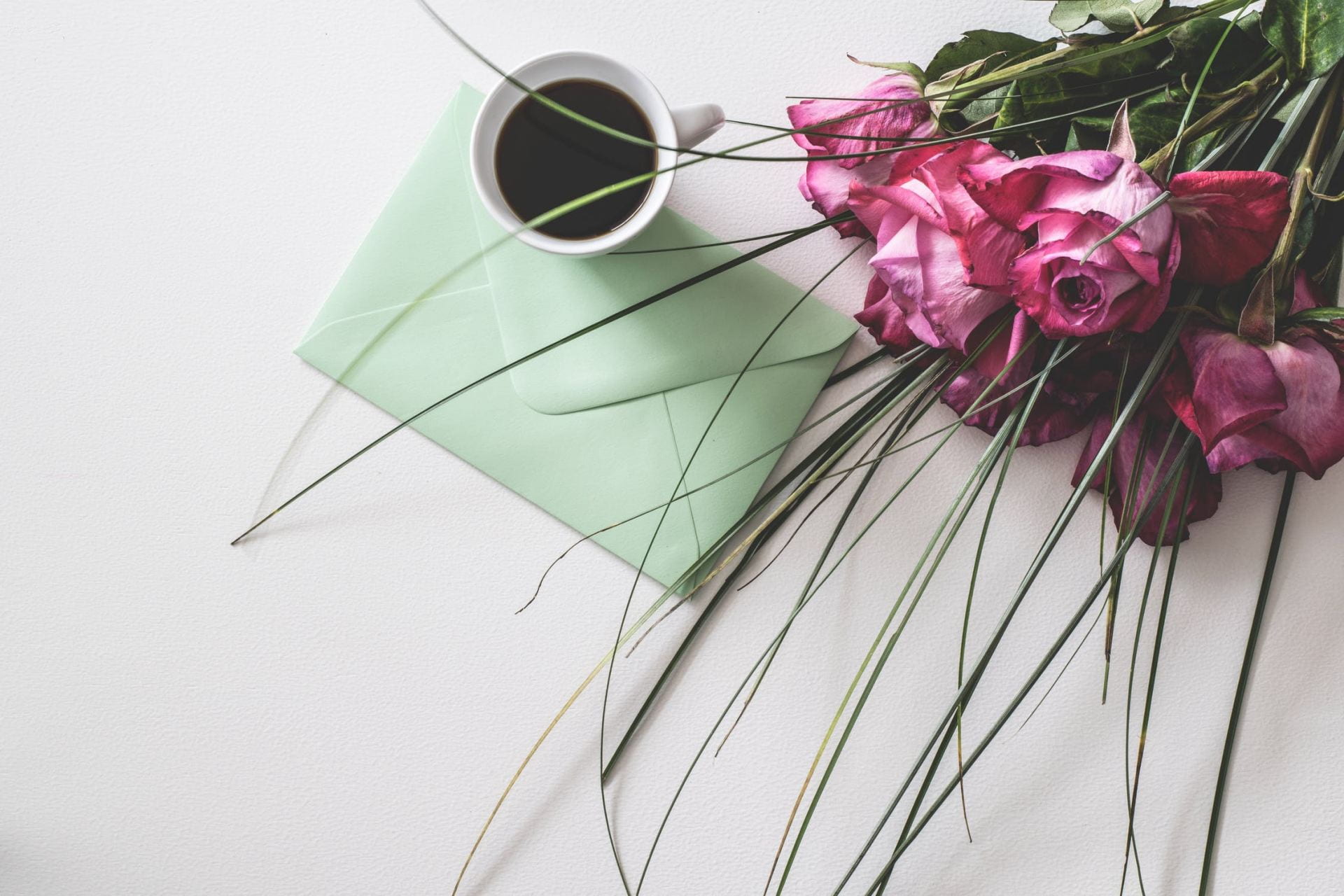 A green letter, a mug of coffee and a bouquet of pink roses on a white surface.