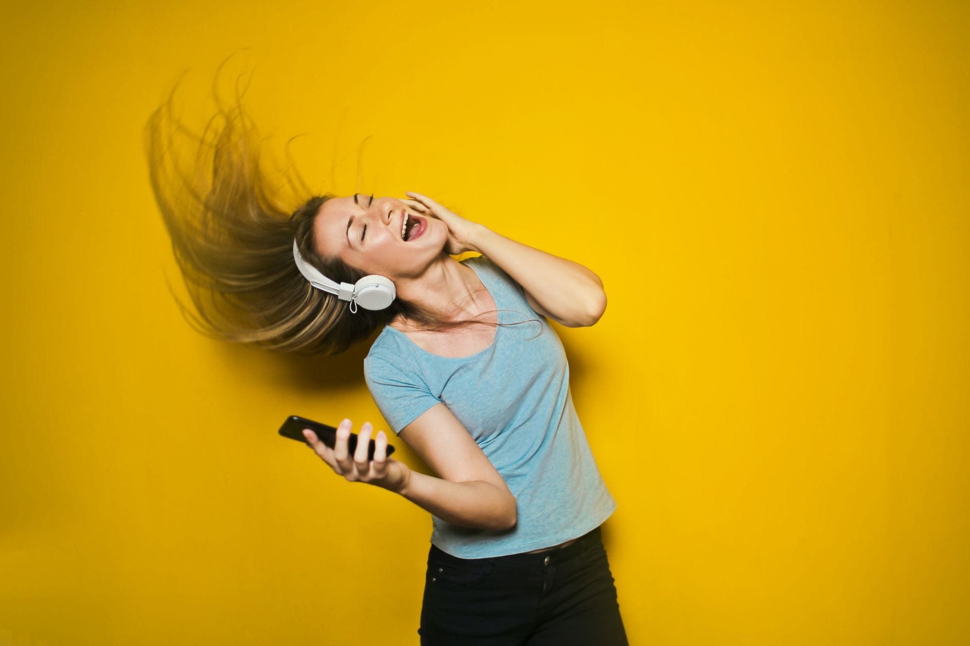 A happy looking woman with headphones on holding a phone on a yellow background.