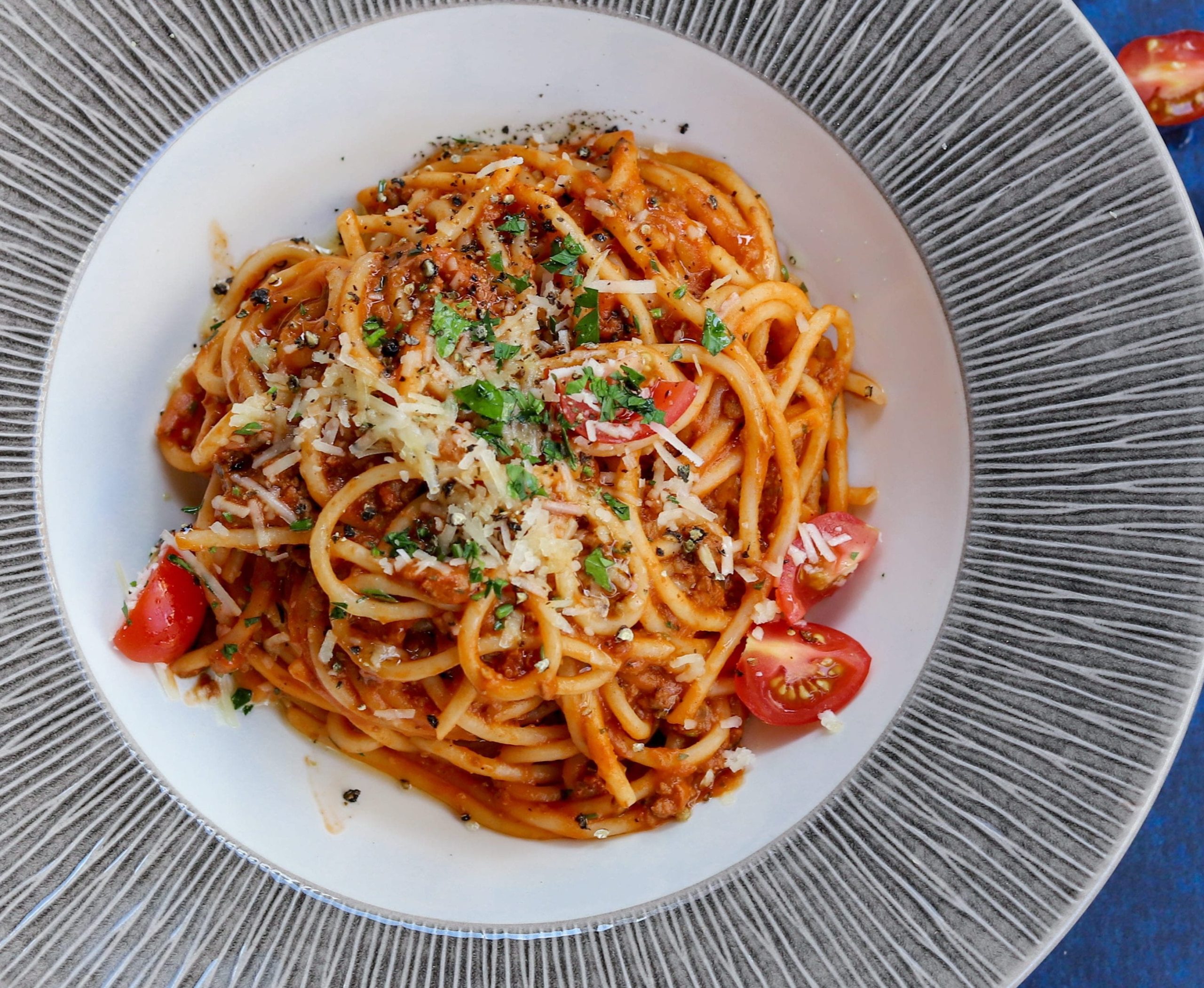A bowl of spaghetti with a tomato sauce. There are sliced tomatoes, chopped green leaves and cheese on top of it.