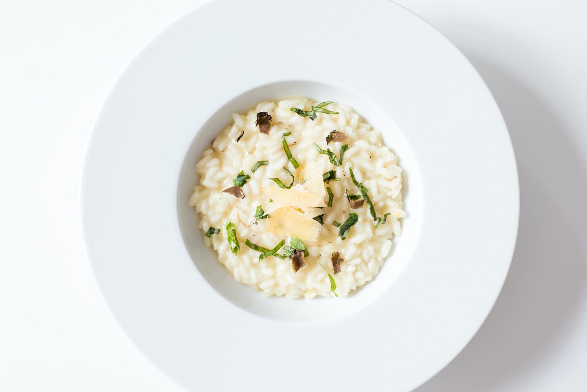A white bowl filled with risotto. On top of the risotto is some green strands and parmesan flakes.