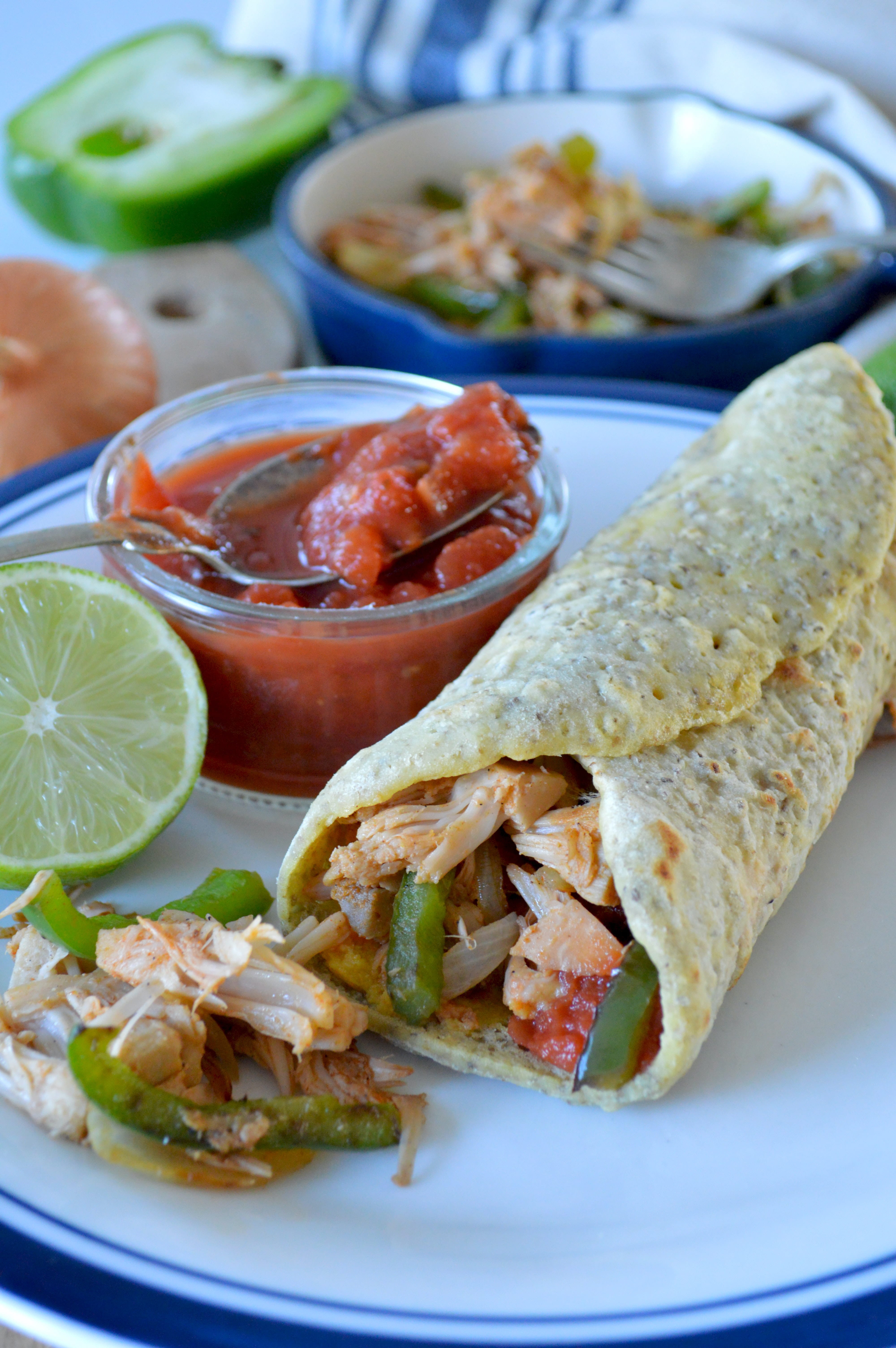 A fajita filled with chicken and vegetables on a plate. There is a pot of salsa and a lime on the plate with it, and a cooking pot of the filling behind it. 