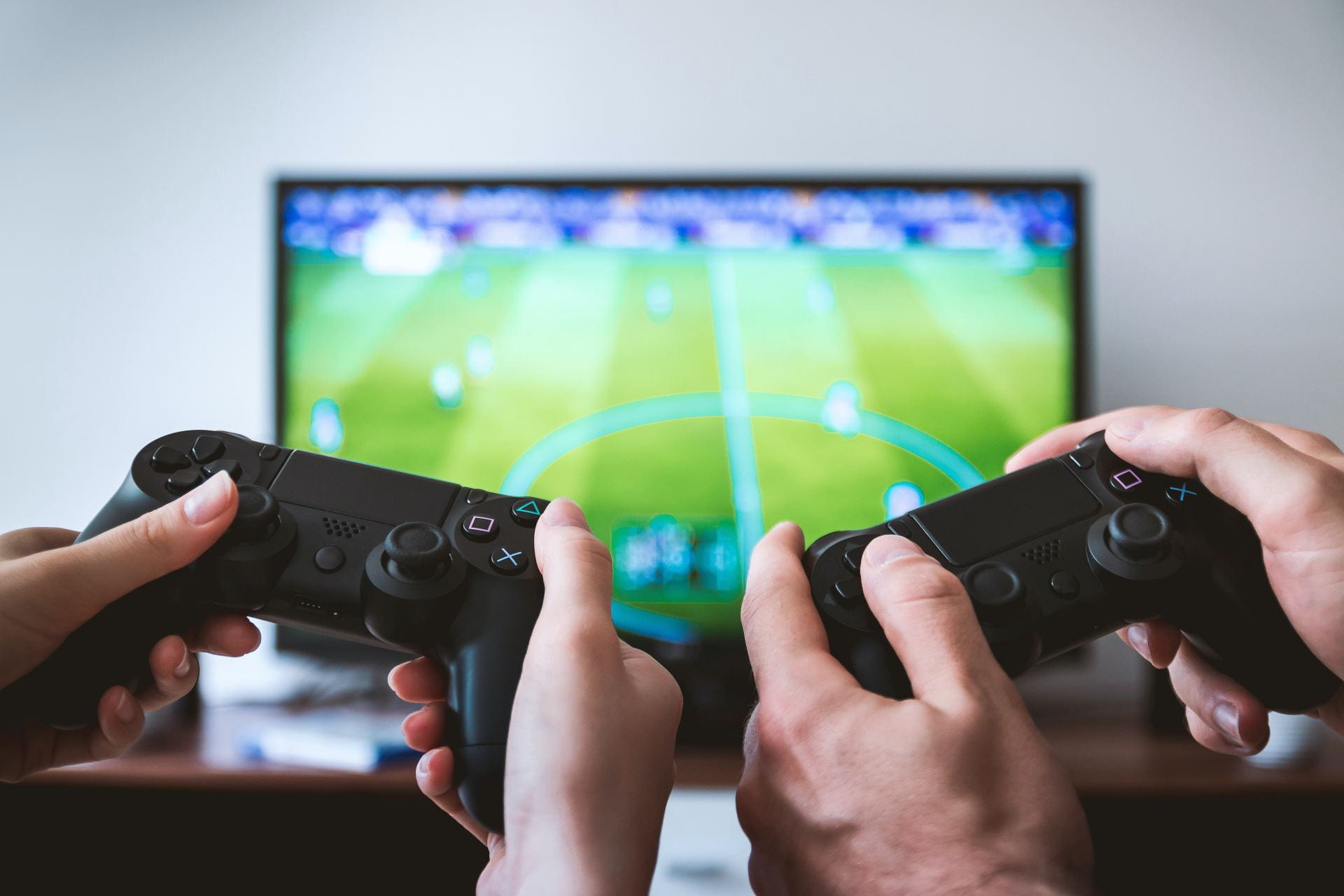 Two pairs of hands holding Playstation remotes. In the background there is a TV out-of-focus with a football game displayed.
