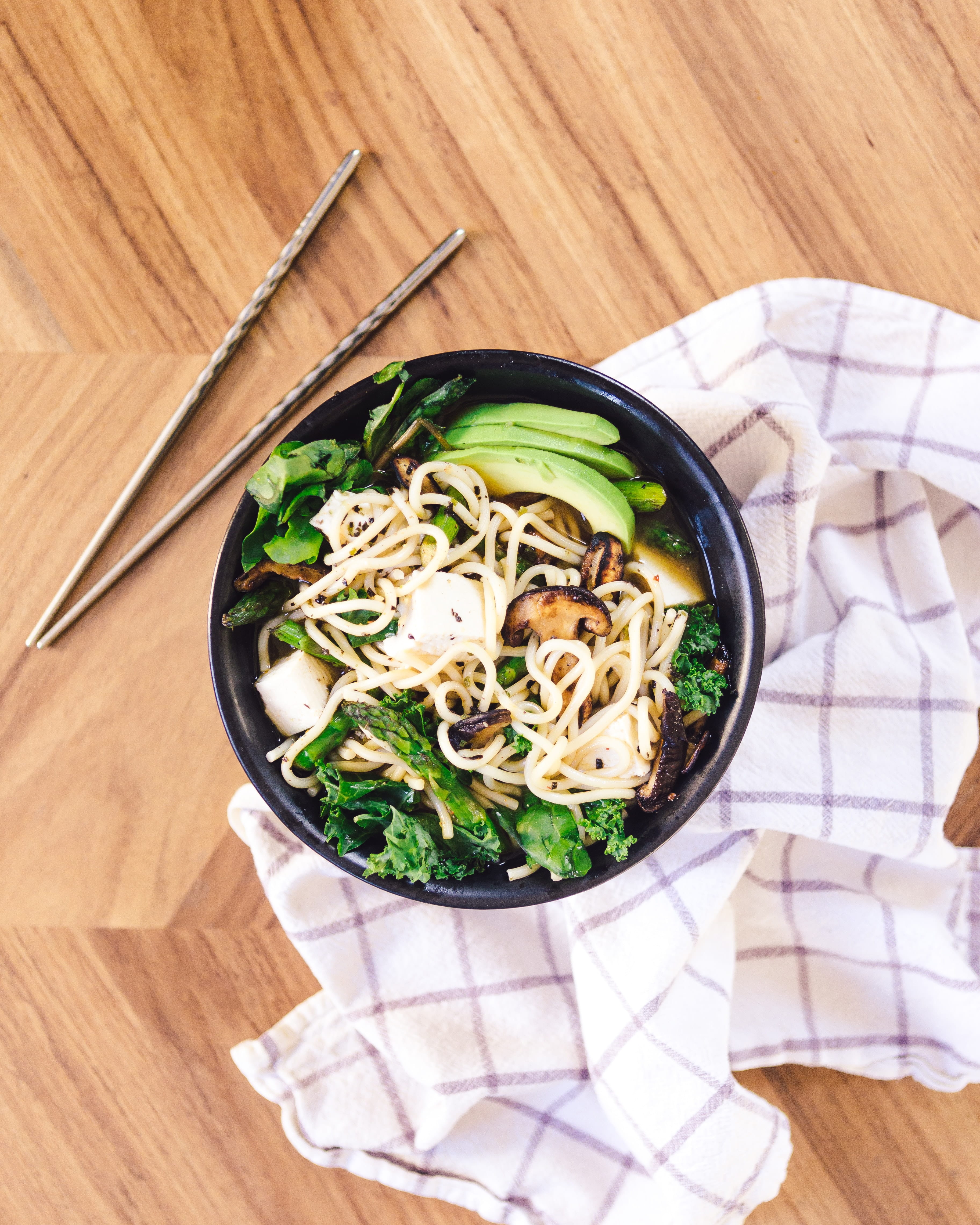 A bowl of ramen with green vegetables and salad leaves. There are chopsticks and a dishcloth to the side of the bowl.
