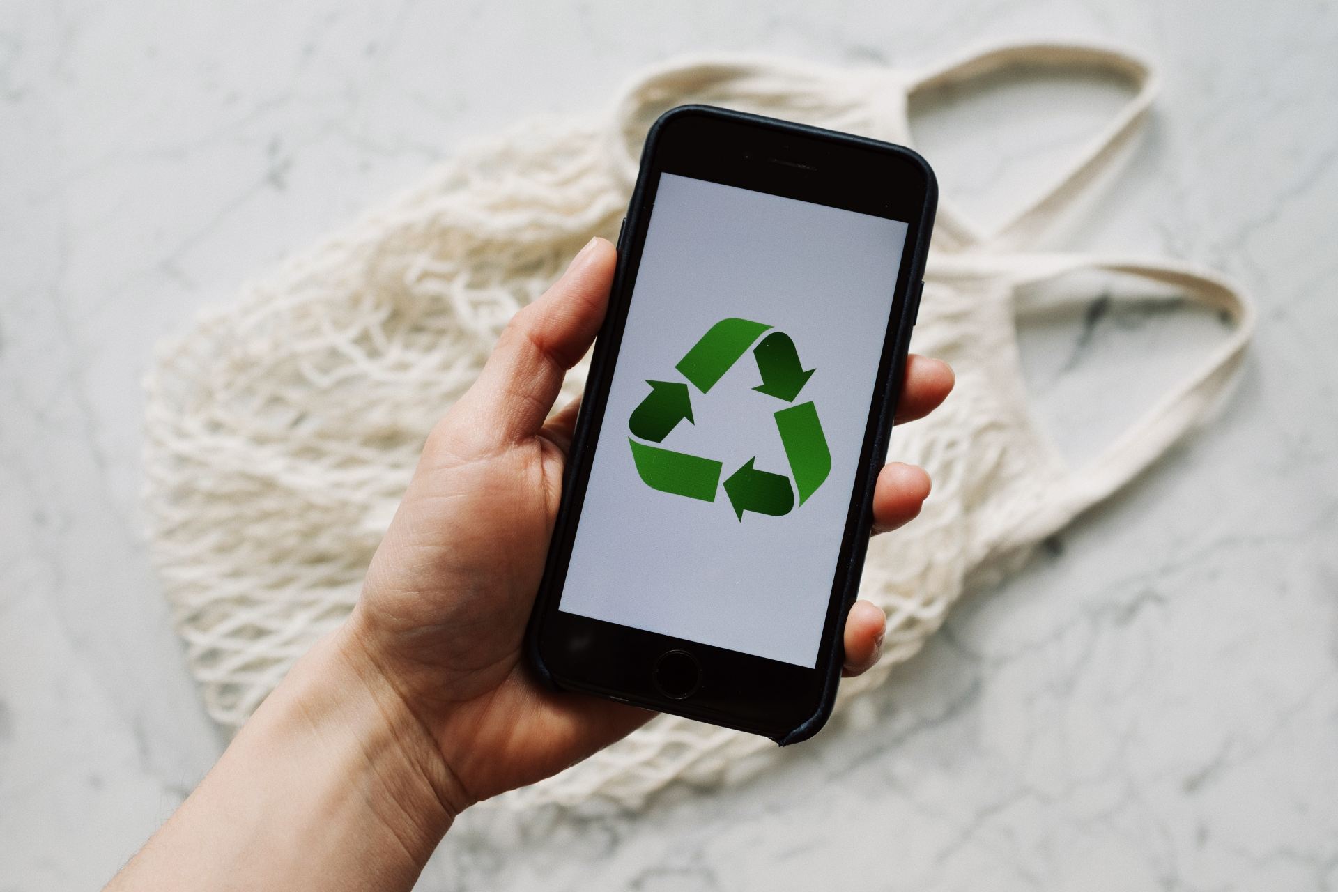 Recycling logo on a phone held in a hand. Below the phone is a reusable bag.