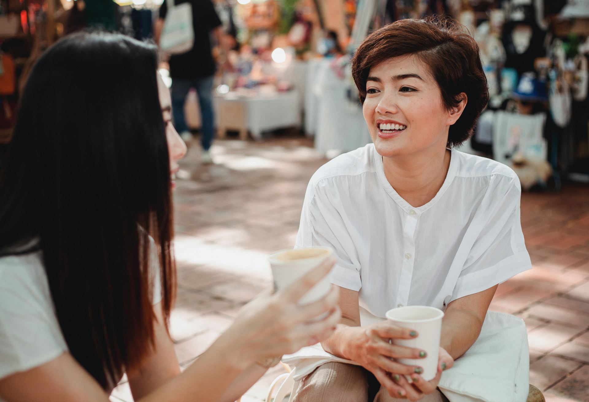 2 women sat down holding paper cups and smiling in a busy street. 