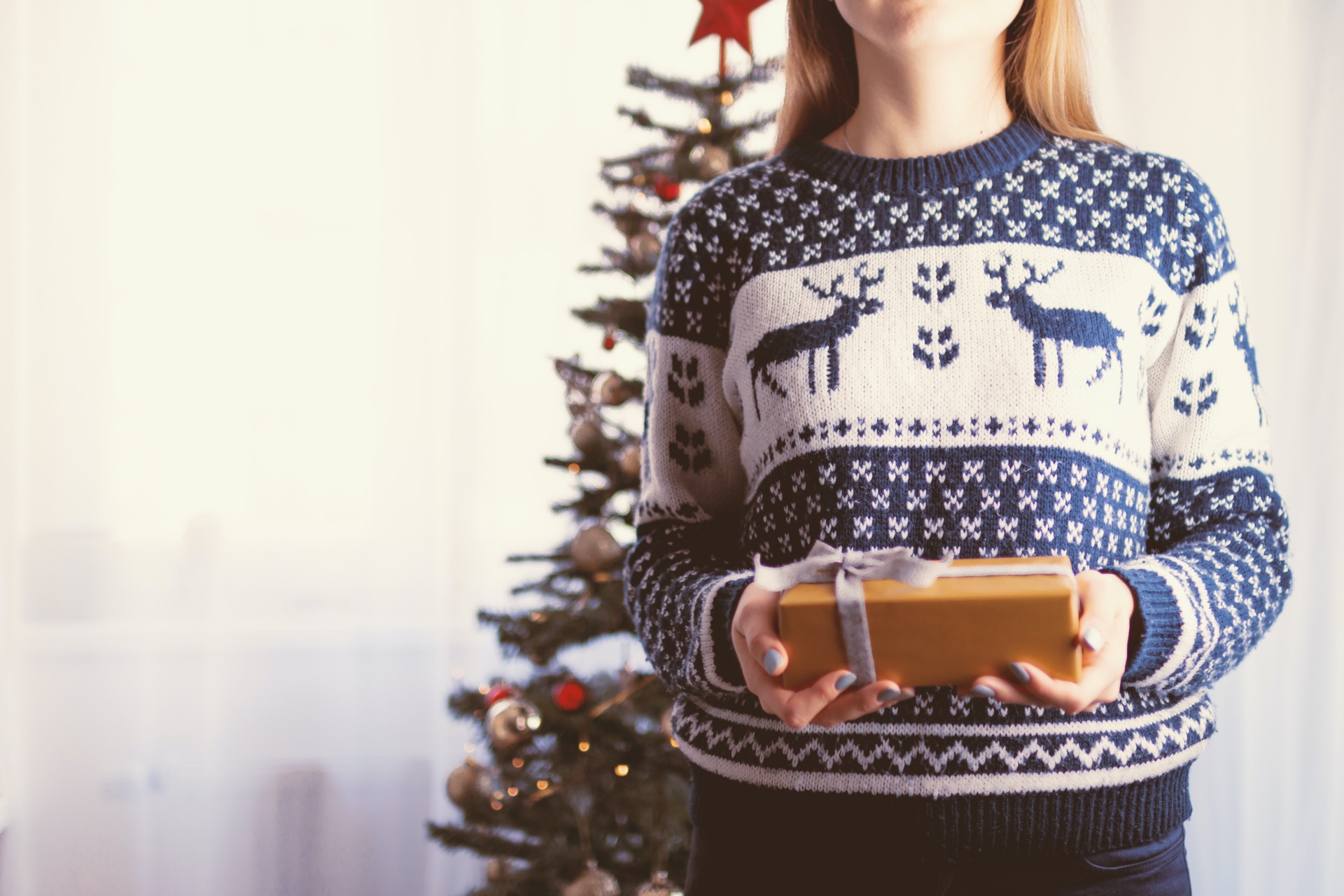 a girl in a Christmas jumper holding a present with a Christmas tree behind her