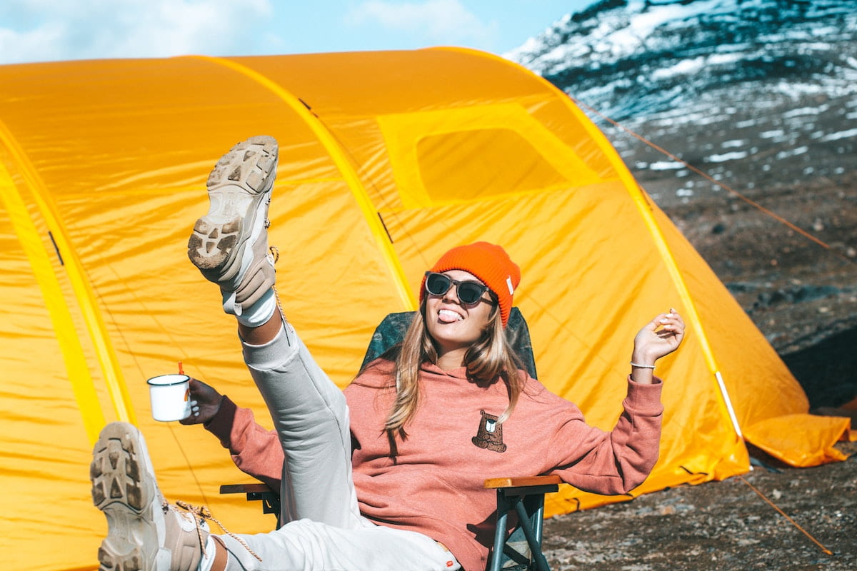 Young woman in a camping chair kicking a foot in the air. Yellow tent behind in sunshine amongst mountains. 