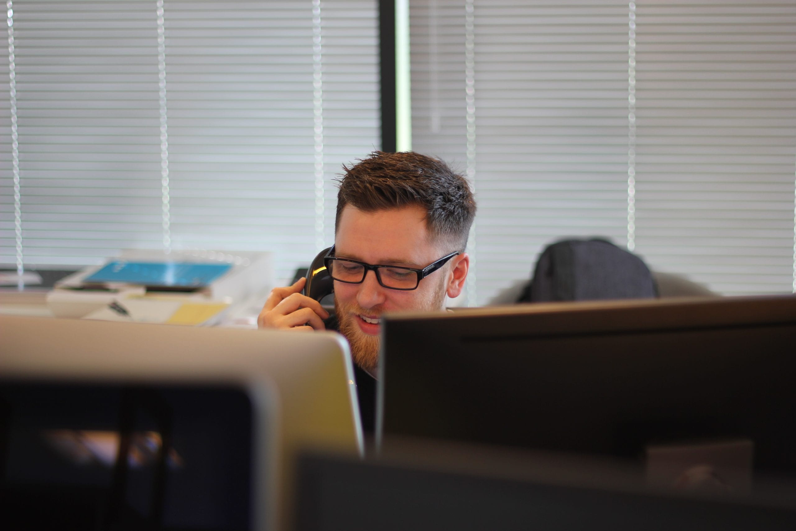 Smiling man wearing glasses sitting behind desk answering telephone. 