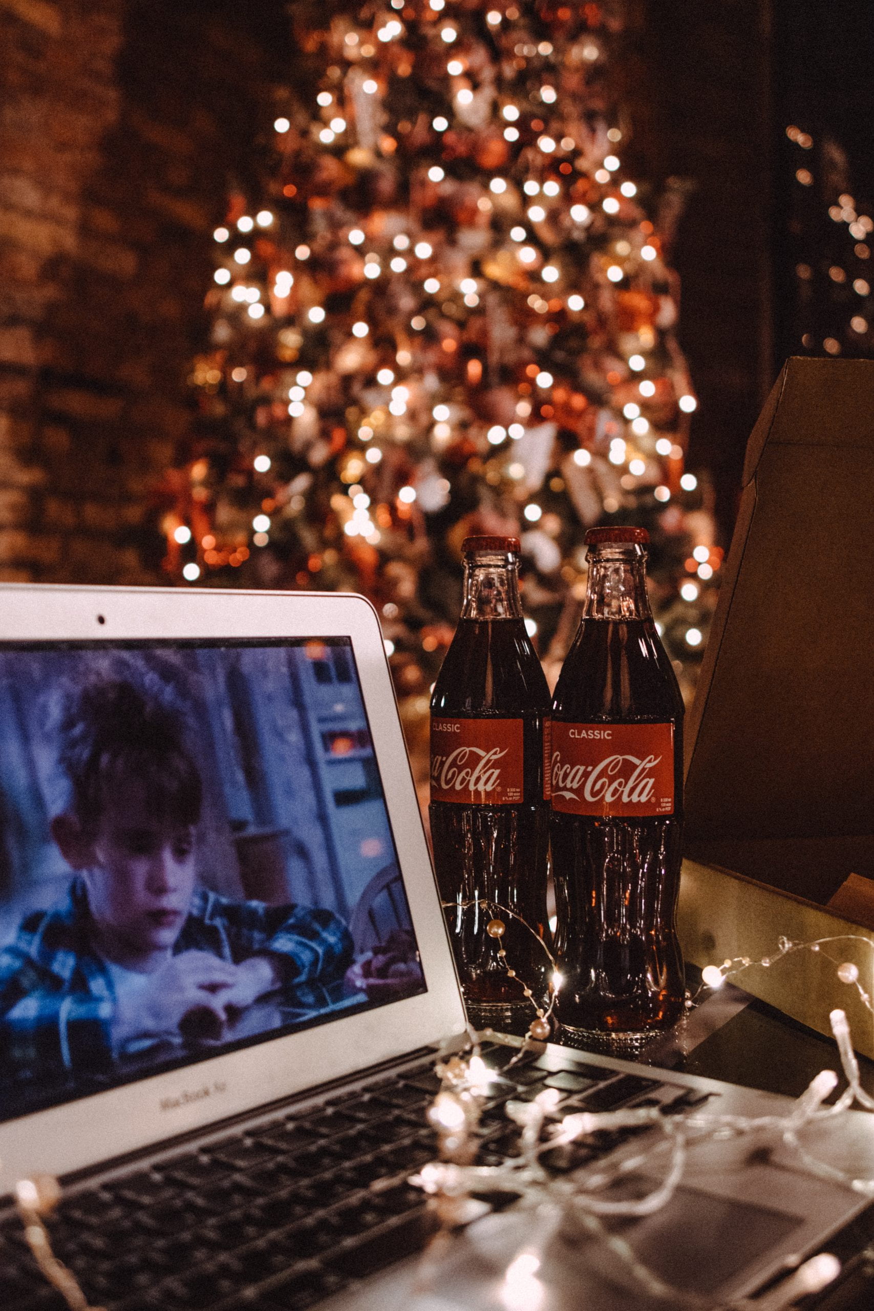 A laptop set up watching the movie Home Alone, Coca Cola (Coke) in the foreground and a brightly lit Christmas tree in the background.