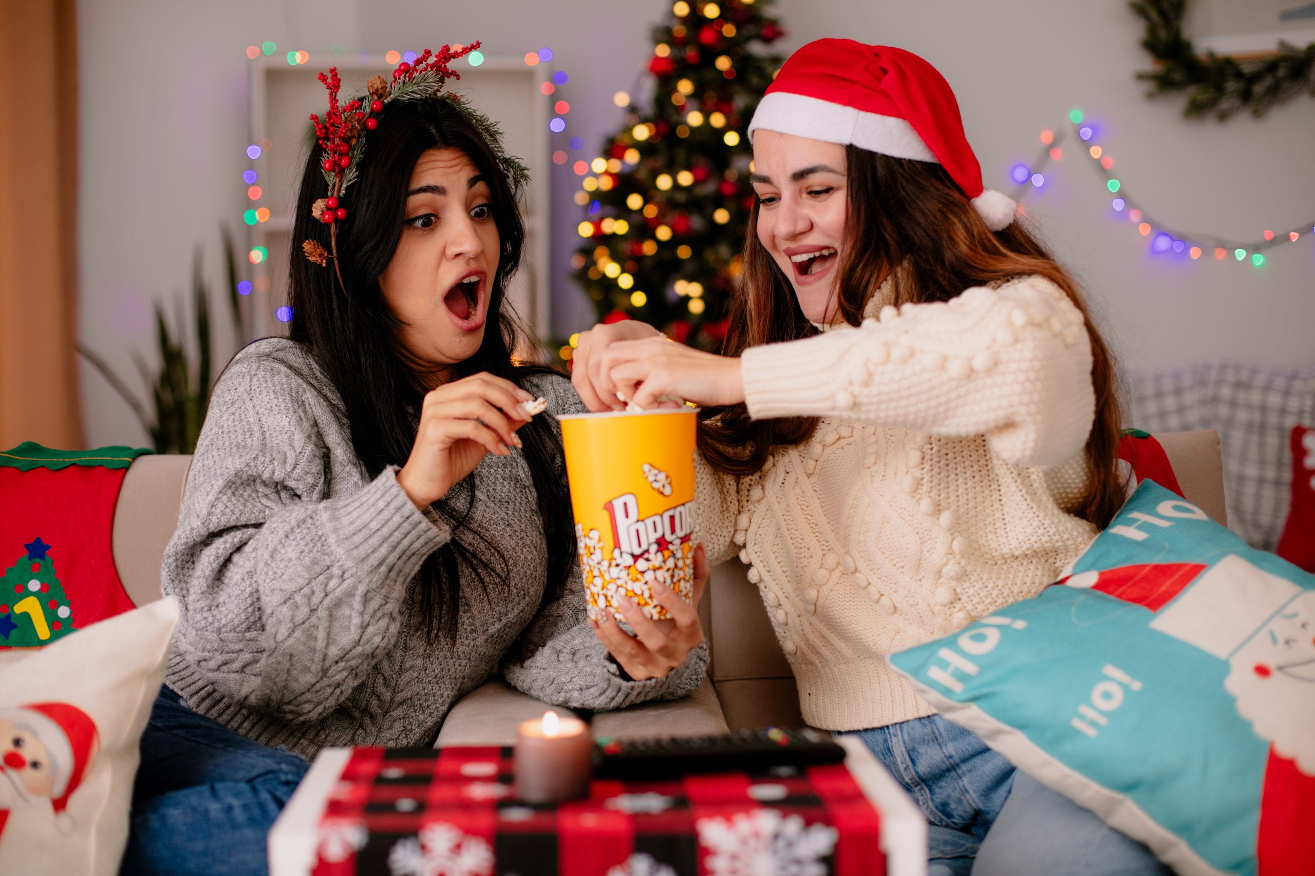 Two women having fun while eating popcorn and watching a movie with Christmas deco in the background.