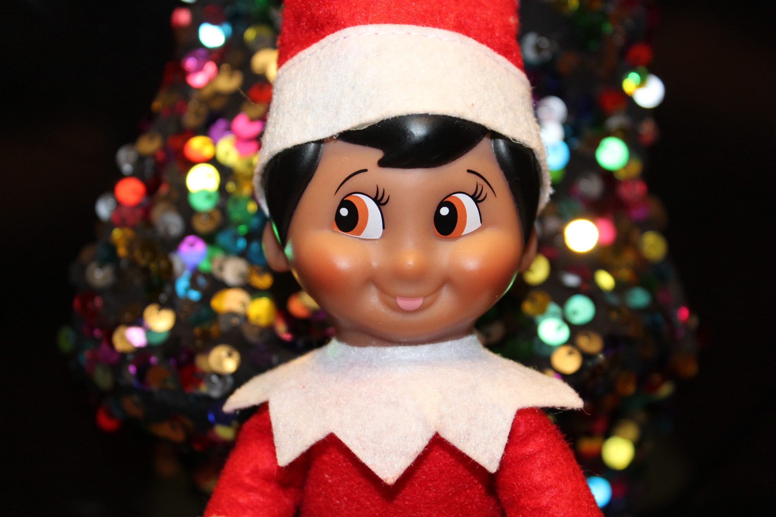 Elf on the shelf sitting in front of a Christmas tree.